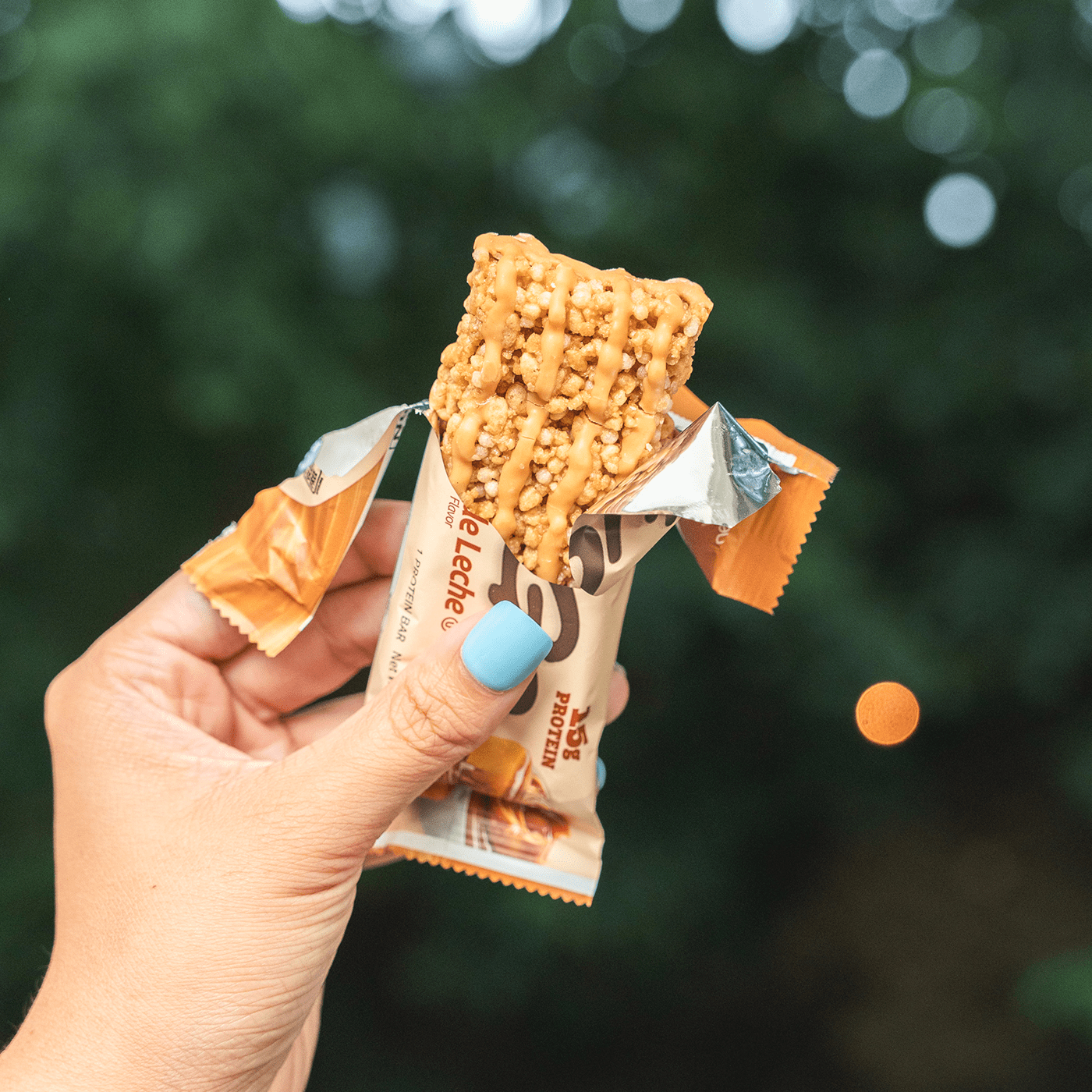 Hand holding a Dulce de Leche protein bar with a blurred green background